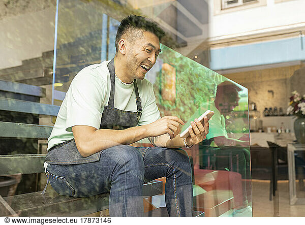 Cheerful man using smart phone sitting on steps seen through glass