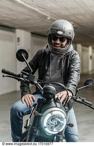 Cheerful man wearing helmet and sunglasses while sitting on motorcycle in parking lot