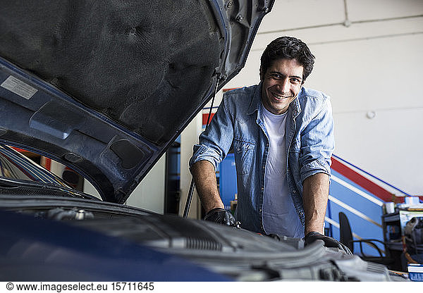 Cheerful man working in car workshop and fixing car engine with tool smiling at camera