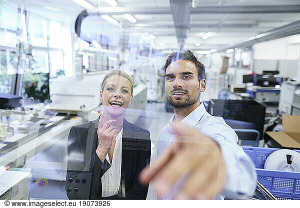Cheerful mature businesswoman standing by businessman pointing at glass interface at factory