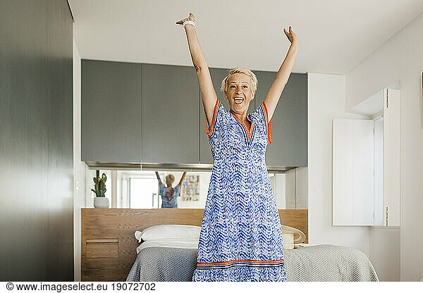 Cheerful mature woman with arms raised standing in bedroom