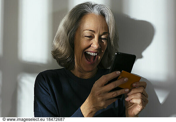 Cheerful mature woman with credit card using smart phone in front of wall