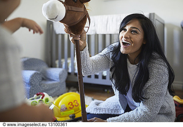 Cheerful mother holding hobby horse while playing with son at home