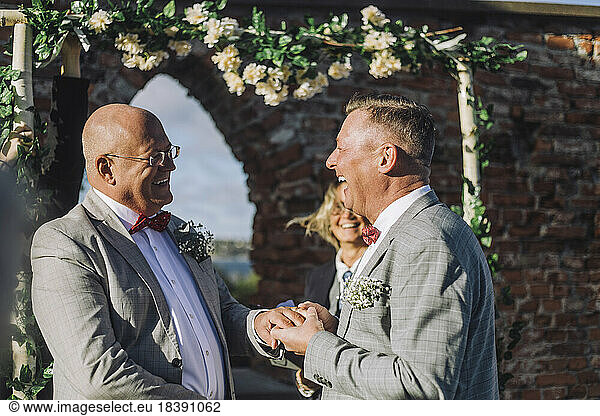 Cheerful newlywed gay couple holding hands by minister at wedding ceremony