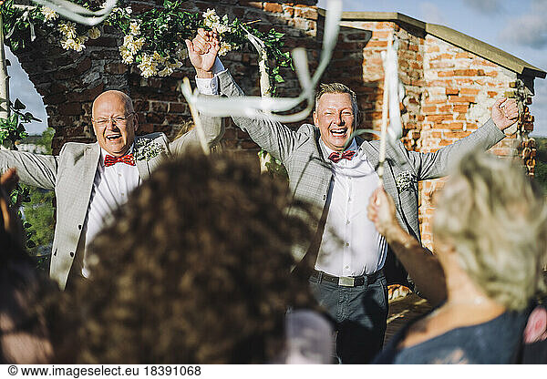 Cheerful newlywed gay couple holding hands while enjoying their wedding ceremony with guests
