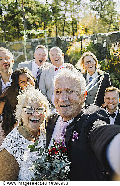 Cheerful newlywed senior couple taking selfie with family and friends at wedding