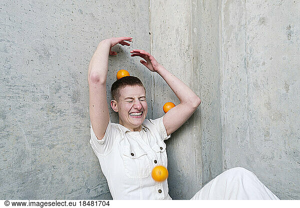 Cheerful non-binary person having fun with oranges in front of wall