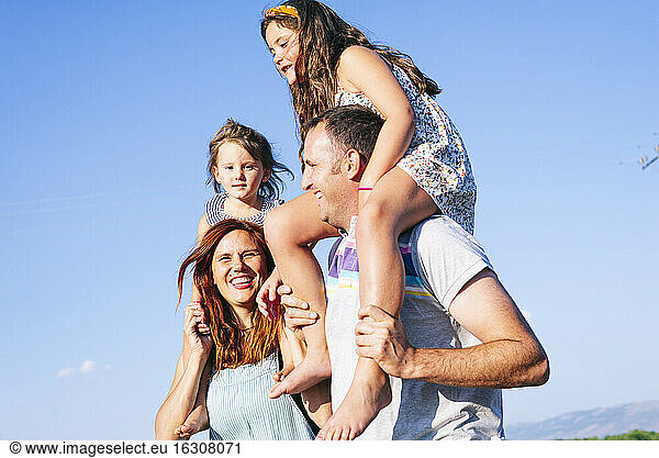 Cheerful parents carrying daughters on shoulders against clear sky during sunny day