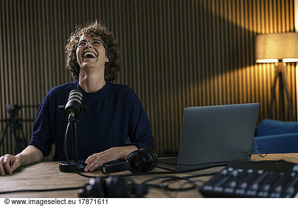 Cheerful presenter with microphone and laptop sitting at desk in radio station