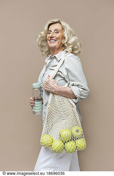 Cheerful senior businesswoman carrying apples in mesh bag against beige background