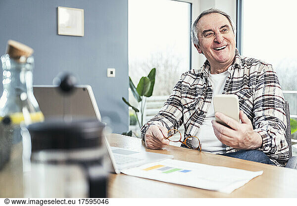 Cheerful senior man holding smart phone and eyeglasses sitting at table