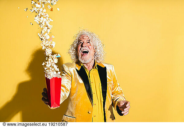 Cheerful senior man throwing popcorns against yellow background