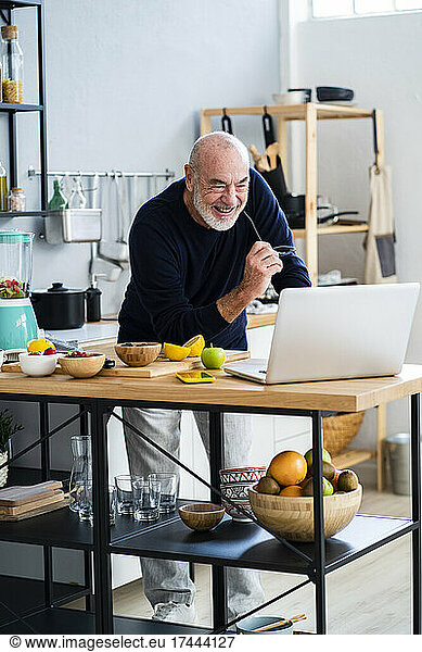 Cheerful senior man using laptop at kitchen counter