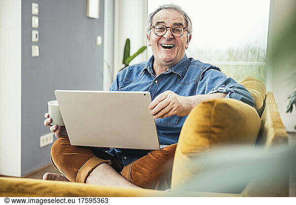 Cheerful senior man with laptop and coffee mug sitting on sofa in living room
