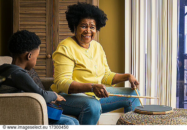 Cheerful senior woman playing drum pad while grandson practicing guitar at home