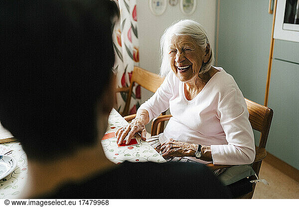 Cheerful senior woman talking with male caregiver while playing cards in kitchen