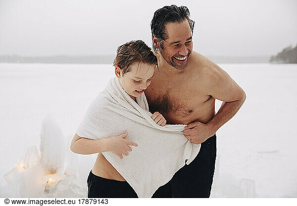 Cheerful shirtless mature man standing with son wearing towel at frozen lake