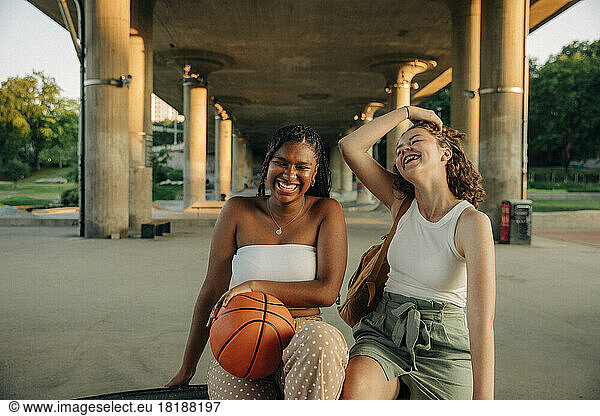 Cheerful teenage girl with basketball sitting by female friend under bridge