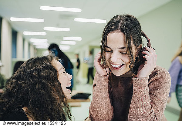 Cheerful teenage girl with headphones sitting with female classmate in corridor at high school