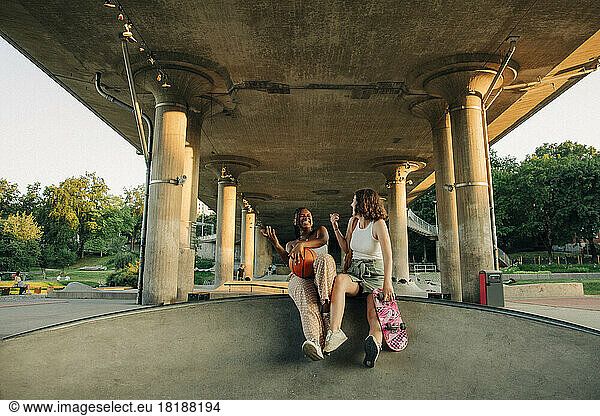 Cheerful teenage girls with basketball and skateboard enjoying under bridge