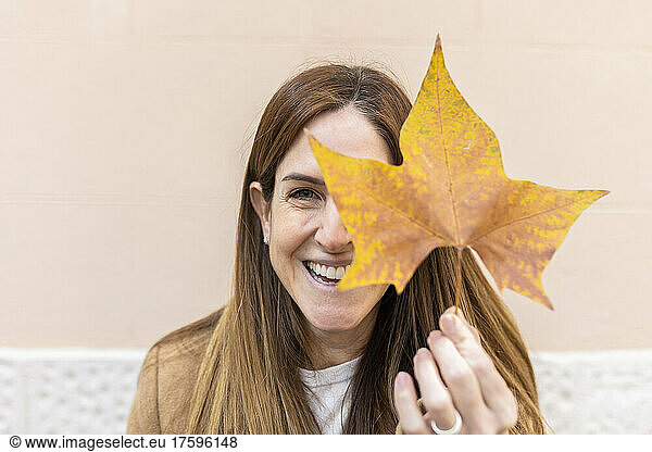 Cheerful woman covering eye with autumn leaf in front of wall