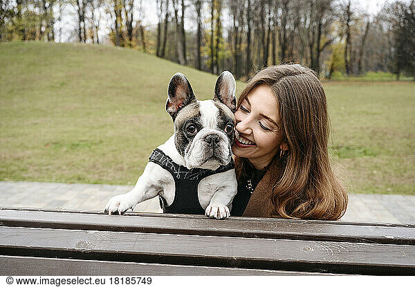 Cheerful woman embracing French bulldog on bench at park