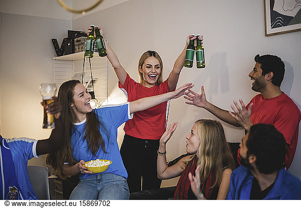 Cheerful woman holding beer bottles standing with friends in living room