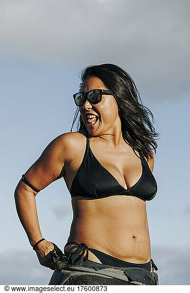 Cheerful woman in sunglasses wearing wetsuit and bikini at beach