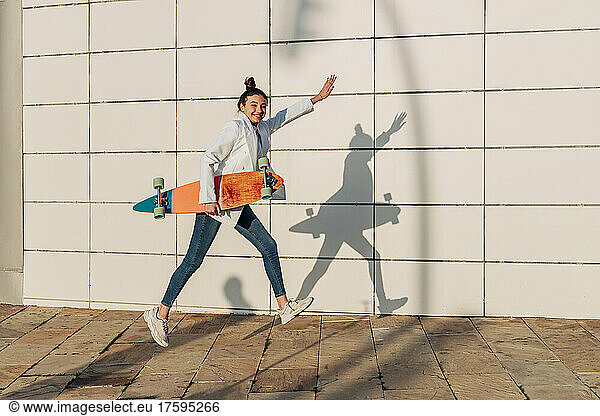 Cheerful woman jumping with skateboard on footpath