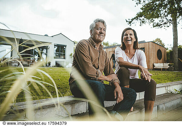 Cheerful woman laughing by senior man sitting on steps at backyard