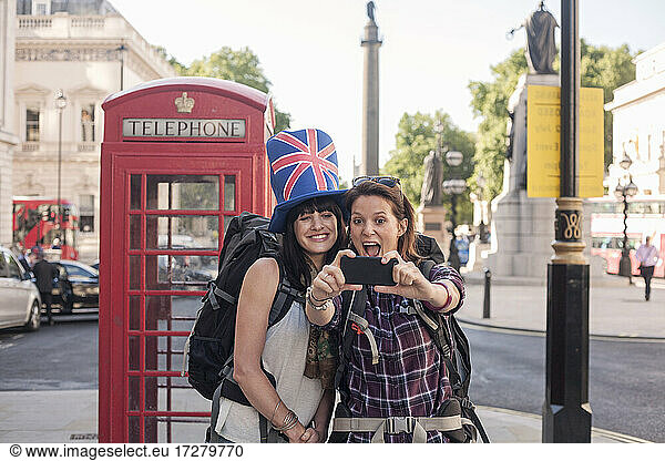 Cheerful woman taking selfie with friend wearing British flag hat against red telephone box in city