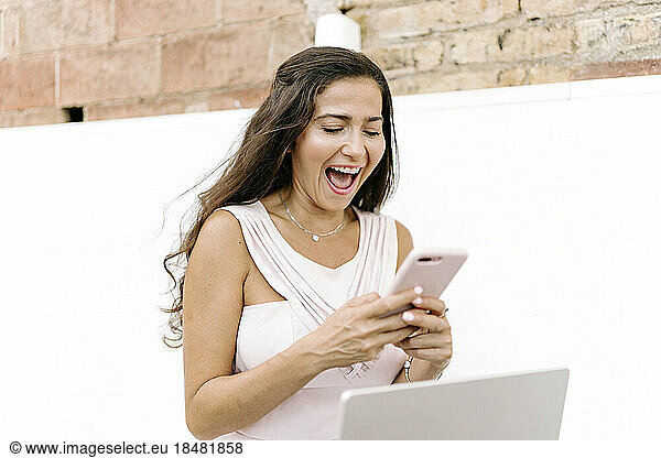 Cheerful woman using smart phone sitting in front of wall