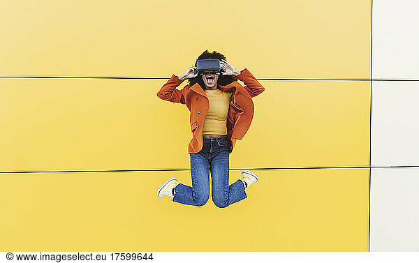 Cheerful woman wearing VR glasses and jumping in front of wall