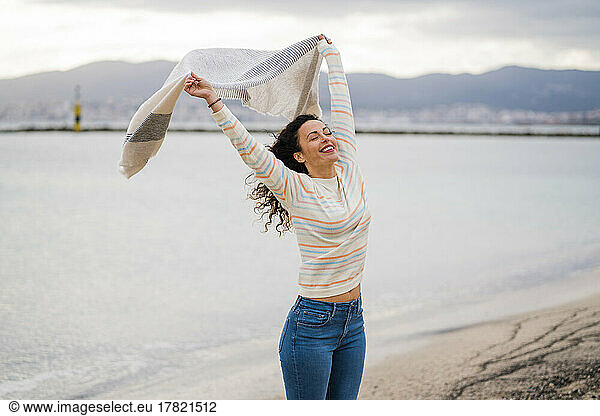 Cheerful woman with arms raised holding scarf at beach