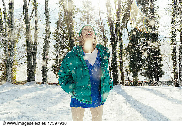 Cheerful woman with hands in pockets enjoying snowfall at forest