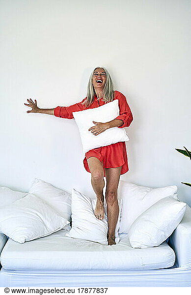 Cheerful woman with pillow standing on sofa