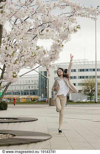 Cheerful young businesswoman jumping by cherry blossom flower tree