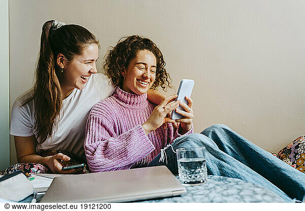 Cheerful young female friends sharing smart phone while sitting at home