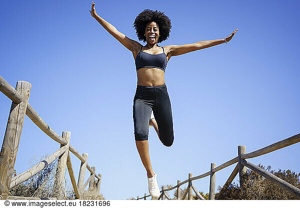 Cheerful young woman jumping over boardwalk under blue sky
