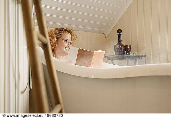 Cheerful young woman lying in bathtub and reading book in daylight