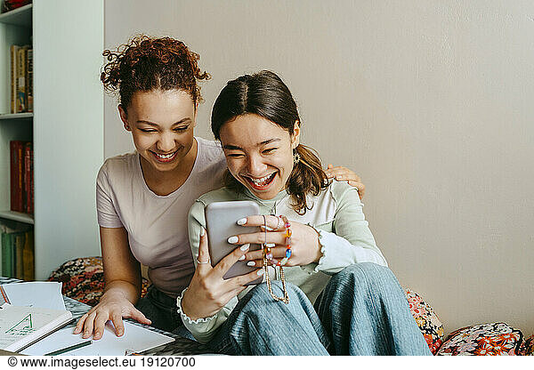 Cheerful young woman sharing smart phone with teenage friend at home