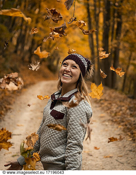Cheerful young woman throwing autumn leaves while enjoying vacation in forest
