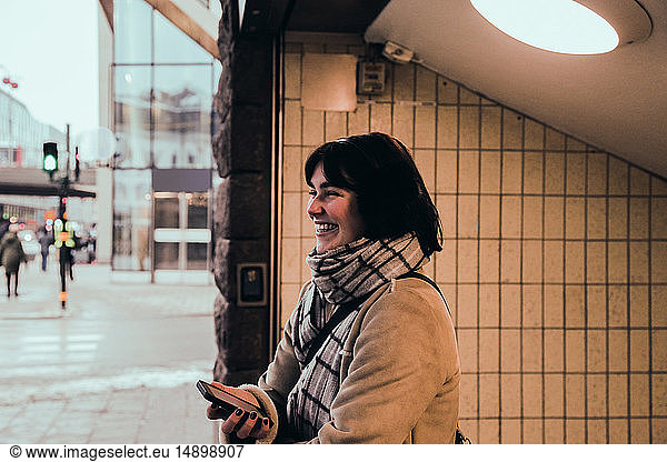 Cheerful young woman wearing warm clothing while standing at subway's entrance in city during winter