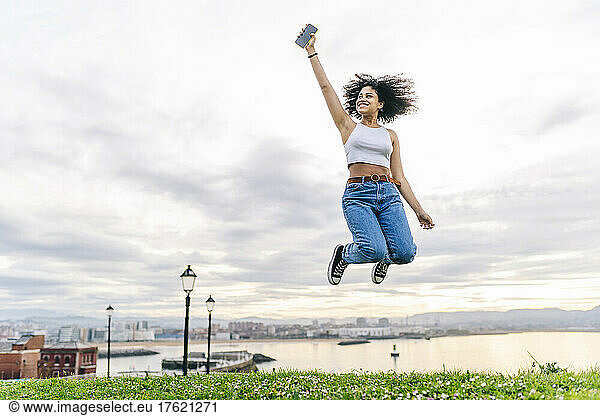 Cheerful young woman with smart phone jumping in nature