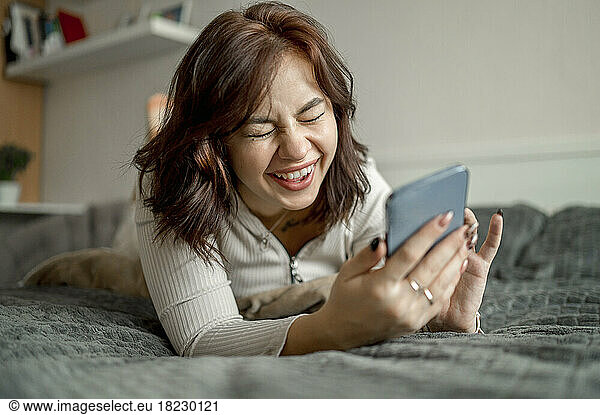 Cheerful young woman with smart phone lying on bed at home