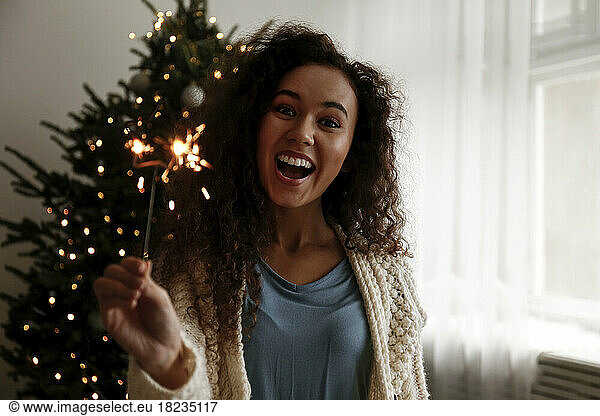 Cheerful young woman with sparkler standing in front of Christmas tree