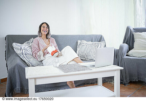 Cheerful young woman with take out food sitting on sofa at home