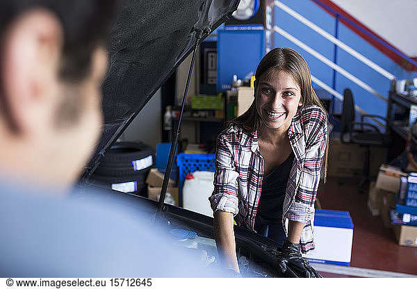 Cheerful young woman working in service station and looking at anonymous man while fixing car engine
