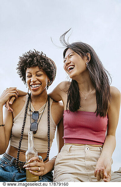 Cheerful young women laughing while sitting against sky