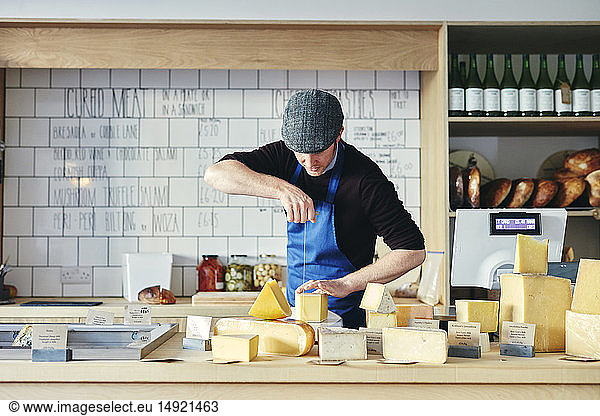 Cheesemonger cutting cheese with cheese wire surrounded by a variety of cheese on counter top